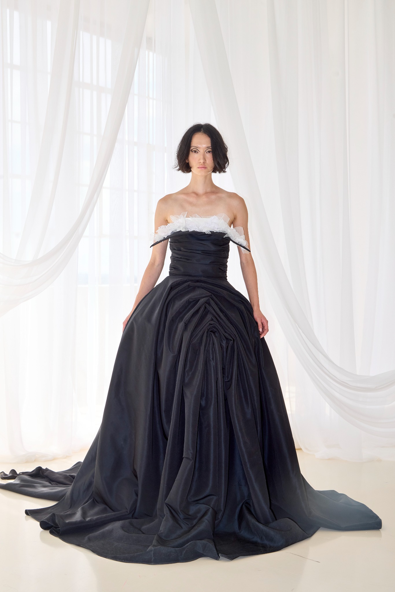 Black full-length gown with a sculptural skirt and white neckline, photographed against white curtains